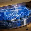 A close-up view of a blue rowing machine component with water splashing around it, set against a wooden background. Produktbild-Vorschau