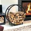 A round black metal firewood rack filled with logs is placed next to a fireplace with a fire burning. Náhľad obrázku produktu