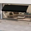 A black awning is extended over a patio area in front of a modern house with large glass windows. Inside the house, a living room with a sectional sofa and coffee table is visible. Náhled obrázku produktu