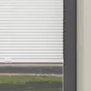 A close-up view of a white roller blind partially covering a window, with a glimpse of grass and pavement outside. Produktbild-Vorschau