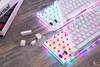 Two white mechanical keyboards with RGB lighting are shown on a wooden surface, with some loose keycaps scattered nearby. A book is partially visible on the left. Produktbild-Vorschau