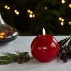 A red spherical LED candle with a flame is placed on a white tablecloth, with pine branches and pinecones in the foreground. In the background, out-of-focus Christmas lights are visible. Produktbild-Vorschau