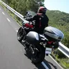 A person riding a motorcycle on a road with trees and hills in the background. Produktbild-Vorschau