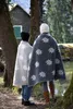 Two people wearing blankets stand in a wooded area near a small cabin. Produktbild-Vorschau