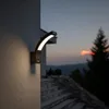 An outdoor LED lamp with a motion sensor is mounted on a textured wall next to a set of stairs, with a twilight sky and trees in the background. Anteprima dell’immagine prodotto