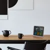 A digital weather station is displayed on a wooden table next to a teapot and a mug, with abstract art on the wall behind. Produktbild-Vorschau