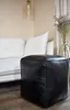 A black leather pouf sits on a wooden floor in front of a white sofa. A vase with dried flowers is visible in the background. Produktbild-Vorschau