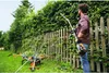 A man is trimming a hedge with a hedge trimmer next to a wooden fence. There is also a lawn mower and other gardening equipment in the background. Produktbild-Vorschau
