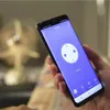 A person is holding a smartphone displaying a smart plug interface, with a blurred fan in the background. Produktbild-Vorschau