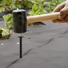 A hand is using a rubber mallet to hammer a black plastic ground stake into gray landscape fabric. Green plants are visible in the background. Náhled obrázku produktu
