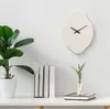 A minimalist wall clock with an organic, asymmetrical shape is displayed on a white wall above a white shelf. The shelf holds a potted plant, a stack of books, and a small vase with greenery. Voir les images du produit