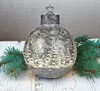 A silver Christmas ornament with a forest and snowflake design sits on a white wooden surface, flanked by pine branches. Produktbild-Vorschau