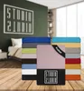 A stack of colorful fitted sheets is displayed in front of a bedroom scene with a bed and a wall graphic. Produktbild-Vorschau