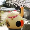 A lucky cat figurine sits in front of a traditional building and cherry blossoms. Produktbild-Vorschau