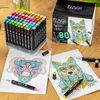 A set of 80 alcohol markers is displayed on a wooden surface, with two coloring pages and some markers laid out in the foreground. Náhled obrázku produktu