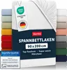 A white fitted sheet is displayed prominently, with a stack of other colored fitted sheets visible on the left and right sides. Several tags and certifications are attached to the sheet. Podgląd zdjęcia produktu