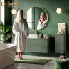 A woman in a white robe stands in a modern bathroom, looking into a round mirror above a green vanity. The bathroom features green tiled walls, a bathtub, and decorative items. Produktbild-Vorschau