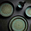A top-down view of a set of ceramic dishes and a mug, arranged on a dark gray surface with scattered dark granules. Náhľad obrázku produktu