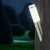 A modern outdoor wall lamp with a motion sensor is mounted on a dark wooden wall, illuminating a garden with manicured bushes at night. Produktbild-Vorschau