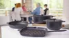 Several black cast iron cookware items are arranged on a white surface in the foreground, with a blurred background showing a family dining at a table. Náhled obrázku produktu
