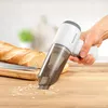 A hand is holding a white and gray handheld vacuum cleaner, cleaning crumbs from a wooden countertop next to a baguette and a knife on a cutting board. Produktbild-Vorschau