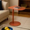 A modern living room scene with a small orange side table next to a textured beige sofa. A bookshelf with a yellow basket is visible in the background. Produktbild-Vorschau