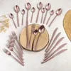 A collection of rose gold cutlery is arranged on a light-colored surface, with some pieces resting on a wooden slice. Podgląd zdjęcia produktu