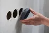A hand is holding a circular gray tool over three circular openings in a gray wall. Produktbild-Vorschau