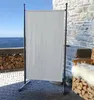 A white fabric screen is set up on a stone patio next to a stack of firewood and a wooden chair with a sheepskin throw. The ocean and sky are visible in the background. Produktbild-Vorschau