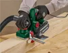 A person wearing gloves is using a green electric planer on a wooden plank. Produktbild-Vorschau
