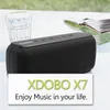 A black portable speaker is placed on a white table next to a glass of water and an open book, with a blurred green background. Produktbild-Vorschau