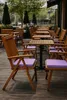 An outdoor seating area with wooden tables and chairs, some with purple cushions. Produktbild-Vorschau