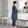 A woman is standing in a bathroom, looking in the mirror and brushing her teeth. A shower stool is visible in the background. Produktbild-Vorschau