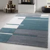 A modern geometric rug with teal, gray, and white sections is laid out on a light gray floor. In the background, a fireplace with logs and a black basket of logs are visible. Voir les images du produit