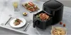 A kitchen counter scene featuring a black air fryer with a whole chicken inside, a plate with a steak and salad, a glass of drink, and a board with lobsters. Produktbild-Vorschau