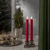 Two red LED candles are lit and placed in candle holders on a surface next to a small potted tree with fairy lights, and a tray of champagne glasses. Produktbild-Vorschau