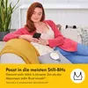 A woman is sitting on a couch, holding a smartphone and using a breast pump. Voir les images du produit
