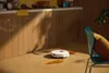 A white robot vacuum cleaner is cleaning up a spill on a wooden floor in a kitchen setting. Sunlight casts shadows on the cabinets. Náhľad obrázku produktu