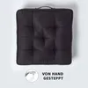 A dark gray square seat cushion is shown from a top-down view against a light gray background. A small circular graphic with a hand sewing is in the bottom left corner, next to text. Náhled obrázku produktu