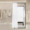 A freestanding changing room with a white curtain and a dark bench inside, set up in a retail environment with clothing racks and garments visible. Voir les images du produit
