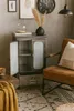A metal cabinet with a glass door is shown in a room with a chair and a rug. Produktbild-Vorschau