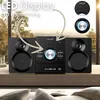 A black mini HiFi system with speakers is shown on a wooden shelf in front of a window with a blurred outdoor view. Produktbild-Vorschau