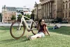 A woman sits on a grassy lawn next to an e-bike, with a large building in the background. Produktbild-Vorschau
