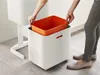 A person is placing an orange inner bin into a white outer bin of a recycling unit. Podgląd zdjęcia produktu