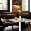 A round aluminum table with a drink and a cup of coffee on it, set in a cafe or restaurant with leather booths and brick walls. Náhled obrázku produktu