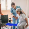 A caregiver assists an elderly woman in transferring from a bed to a wheelchair using a blue transfer board. Produktbild-Vorschau