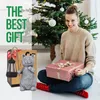 A woman is sitting on the floor in front of a Christmas tree, holding a gift. There are also decorative items like a cat figurine and a lantern in the foreground. Produktbild-Vorschau