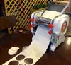 A dough sheeter machine is shown on a wooden table, producing a long sheet of dough with circular cutouts. There are also pre-cut dough circles and a small container nearby. Náhled obrázku produktu