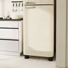 A close-up view of a cream-colored retro refrigerator in a kitchen setting. Part of a white cabinet with drawers is visible to the left, and a marble-patterned floor is shown below. Kitchen utensils hang on a rack above the cabinet. Produktbild-Vorschau