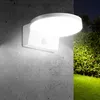 A white LED wall lamp with a motion sensor is mounted on a concrete wall, illuminating a green lawn and foliage at night. Voir les images du produit